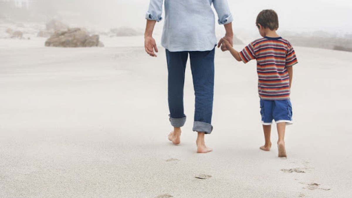 Father and son holding hands while walking on sand at beach – fr