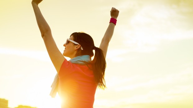 Happy successful sportswoman raising arms to the sky on golden back lighting sunset summer. Fitness athlete with arms up celebrating goals after sport exercising and working out outdoors. Copy space.