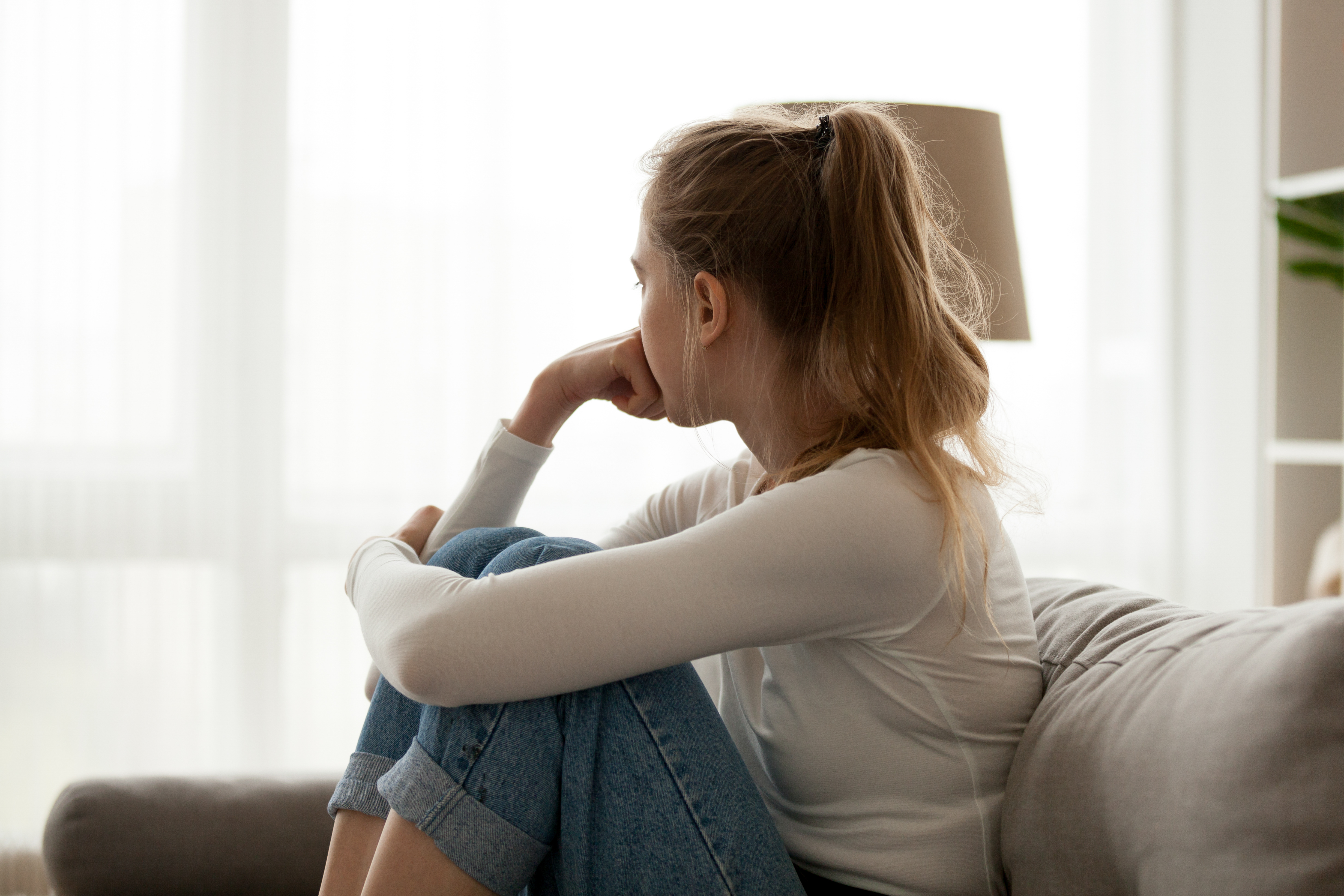 Vista lateral de una joven mirando a la ventana sentada en el sofá en casa. Frustrada mujer confusa se siente infeliz problema en la vida personal